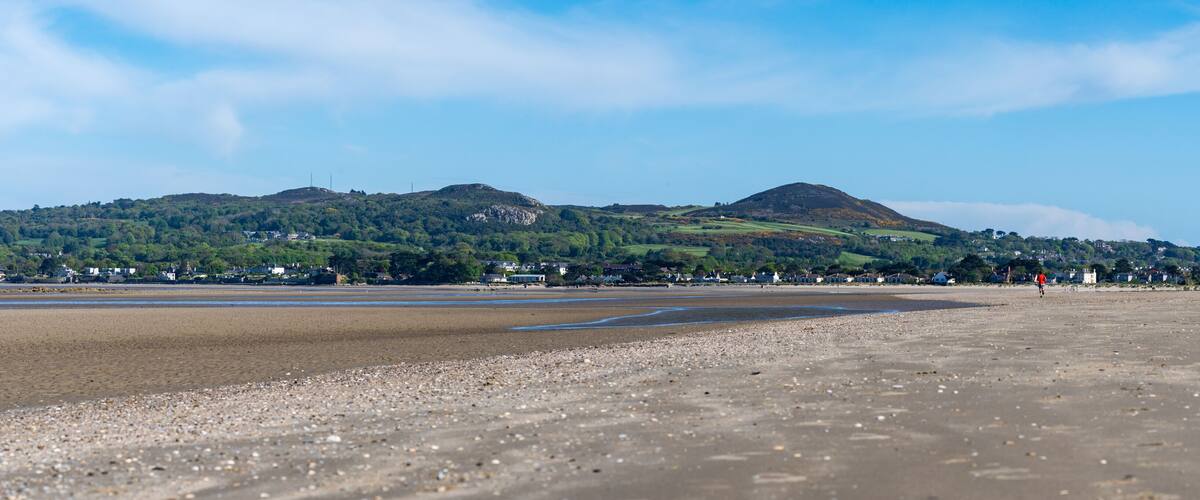 A beach with pebbles, a distant hill with houses, and a clear sky.