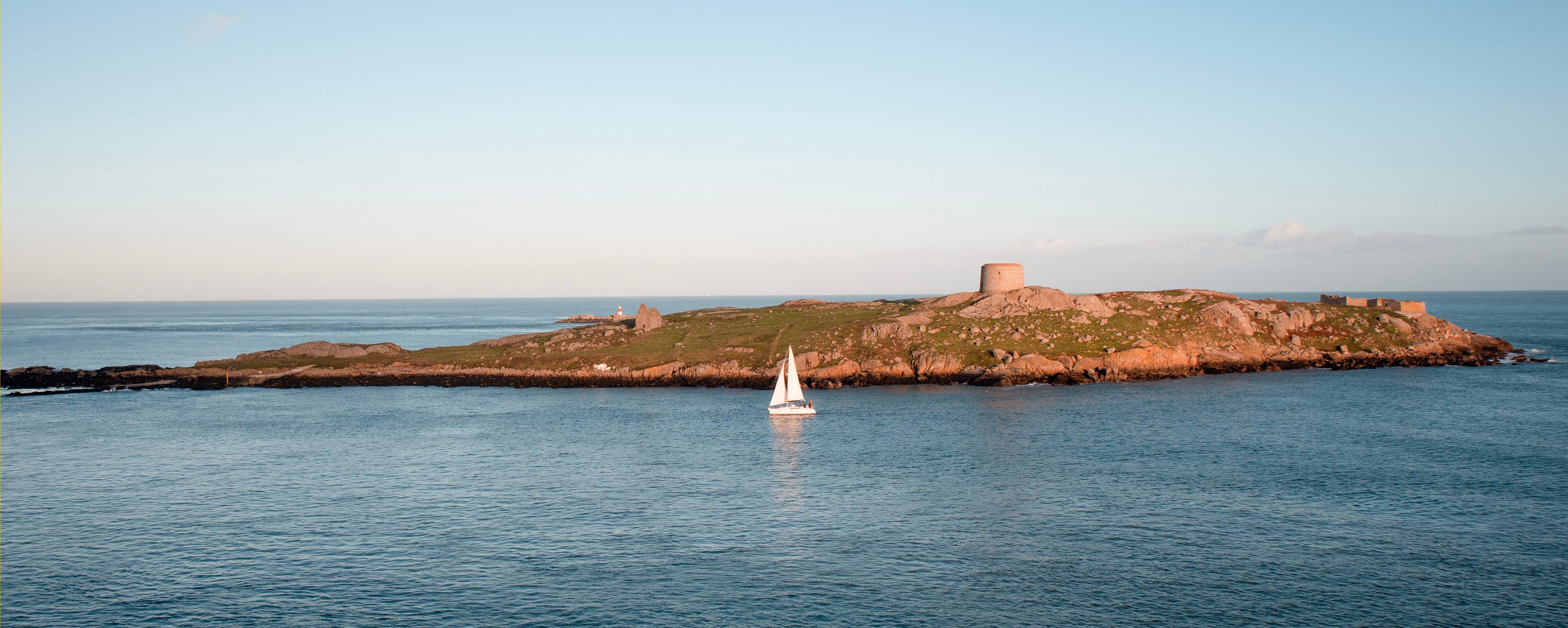 A sailboat on calm waters near a rocky island with a prominent tower.