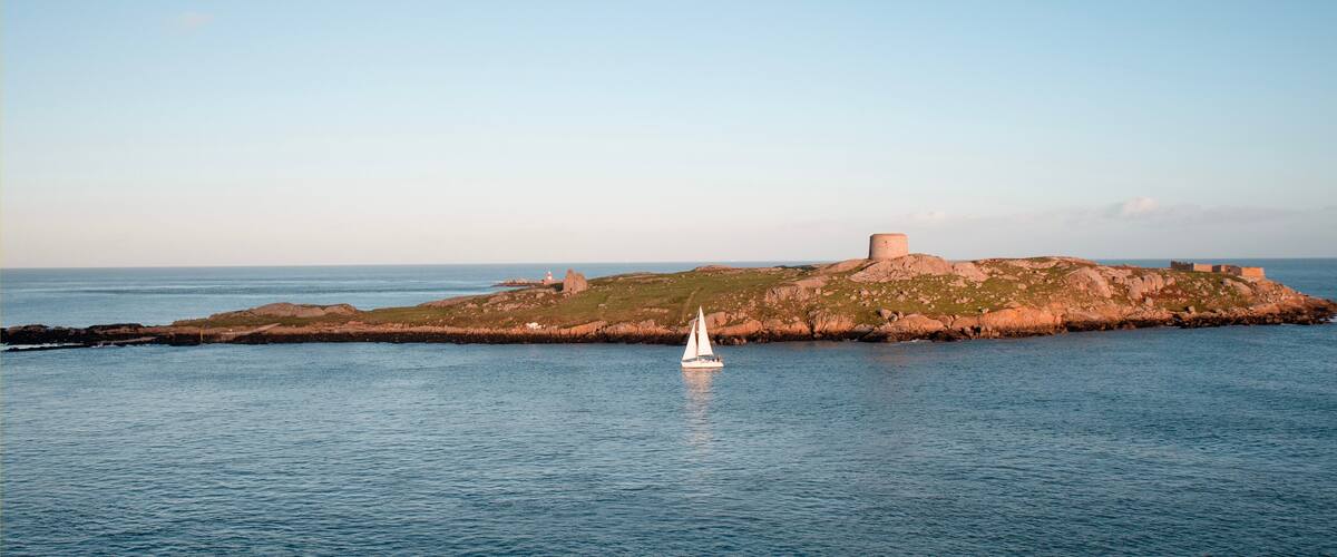 A sailboat on calm waters near a rocky island with a prominent tower.