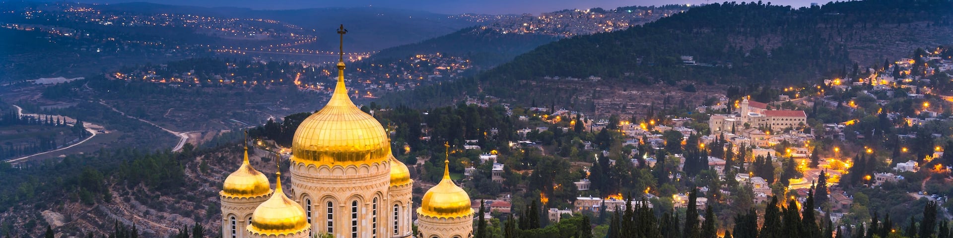 A cathedral with golden domes, surrounded by a mountainous landscape and a lit-up town in the background.