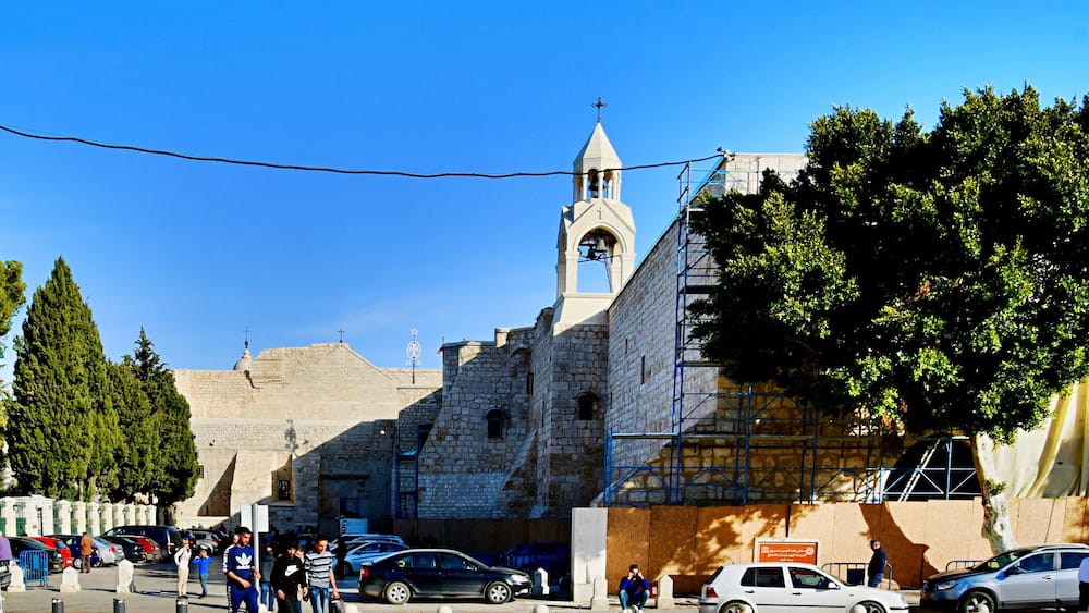 A historic stone building with a bell tower, surrounded by parked cars and pedestrians.