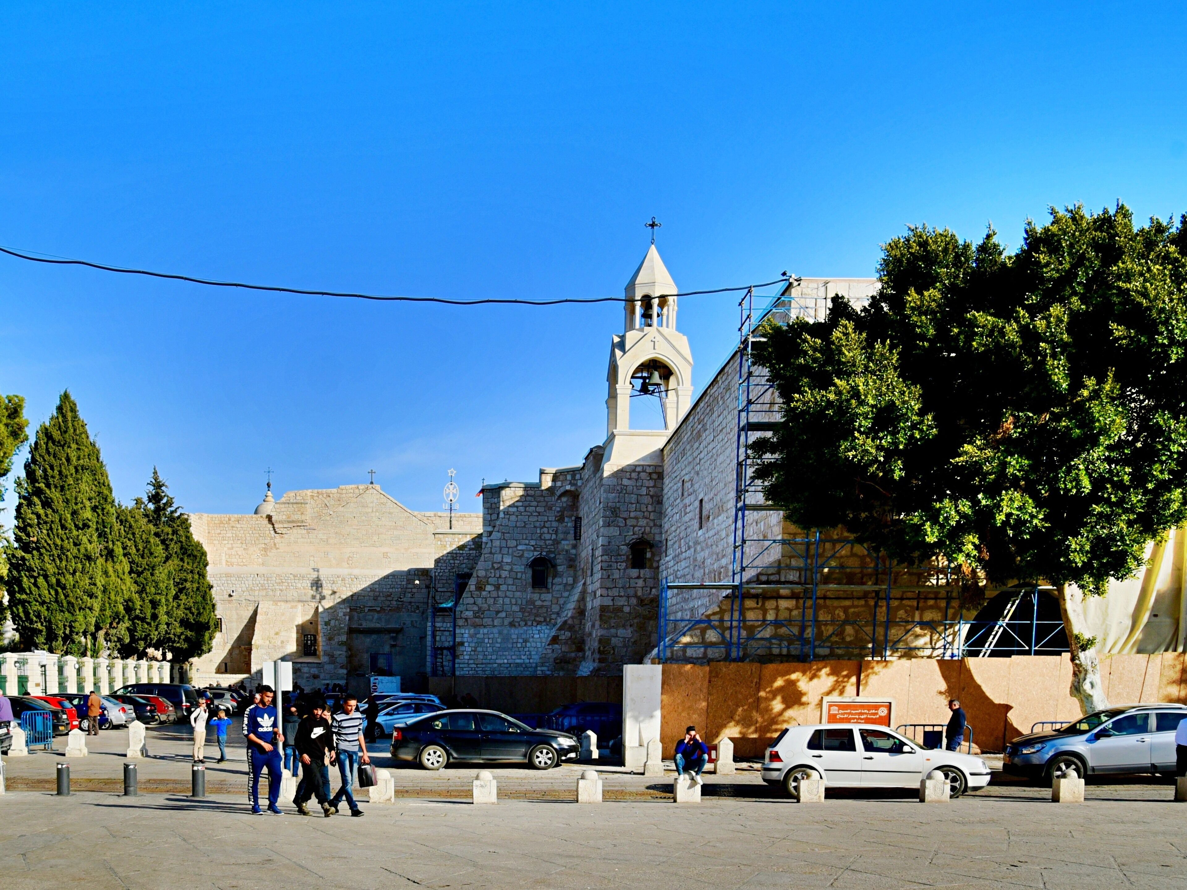 A historic stone building with a bell tower, surrounded by parked cars and pedestrians.