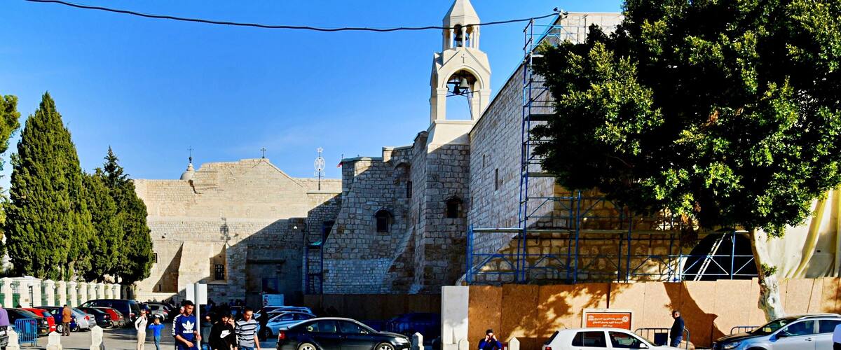 A historic stone building with a bell tower, surrounded by parked cars and pedestrians.