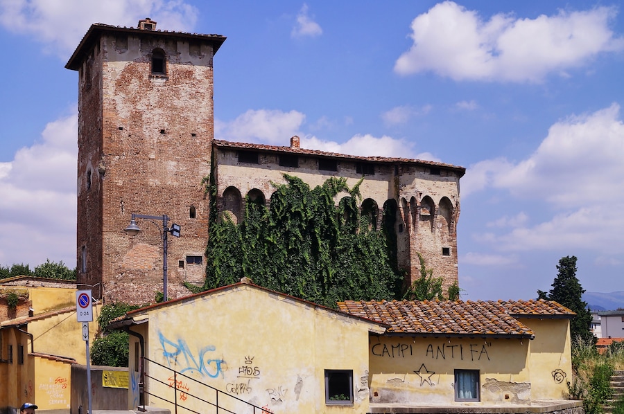Un edificio storico con un'alta torre e finestre ad arco, immerso nel verde sotto un cielo sereno.