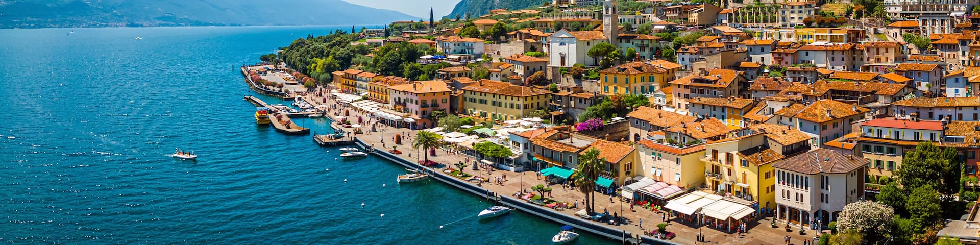 A coastal town with terracotta-roofed buildings, a marina, and mountains in the background.