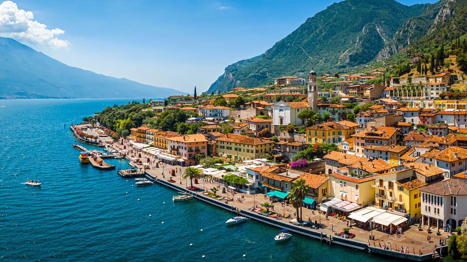A coastal town with terracotta-roofed buildings, a marina, and mountains in the background.