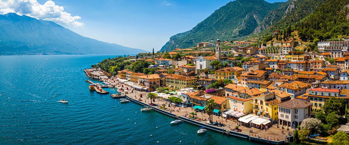 A coastal town with terracotta-roofed buildings, a marina, and mountains in the background.
