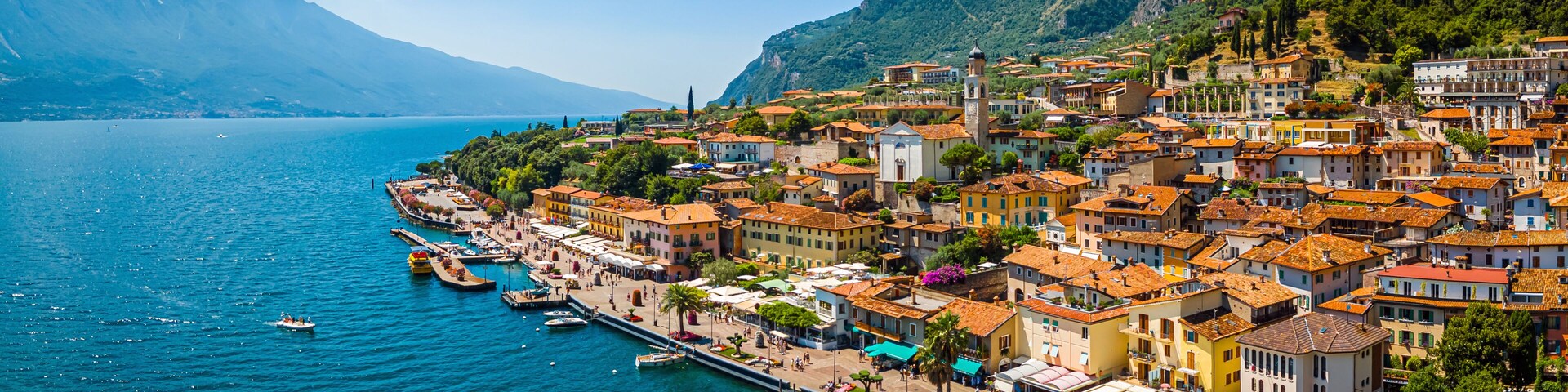 A coastal town with terracotta-roofed buildings, a marina, and mountains in the background.