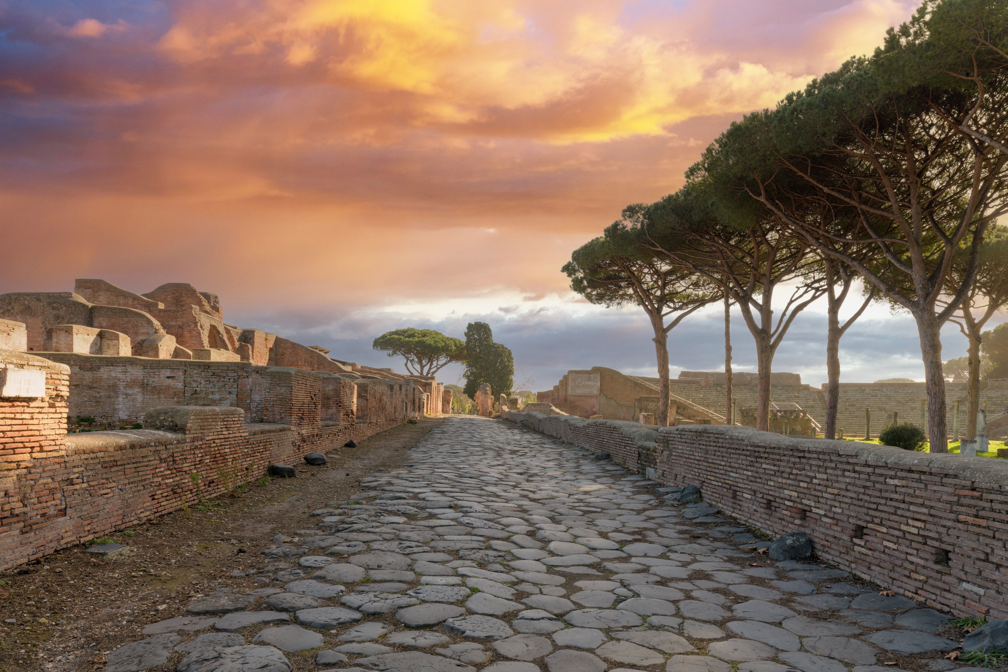 An ancient stone-paved pathway flanked by brick walls and trees, leading towards ruins under a dramatic sky.