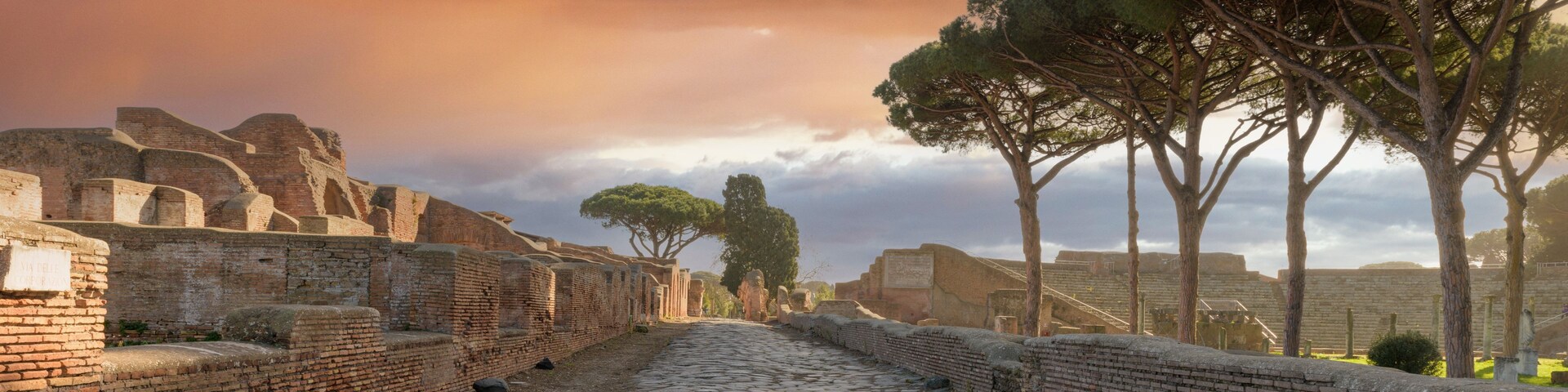 Un antiguo sendero empedrado, flanqueado por muros de ladrillo y árboles, que conduce a unas ruinas bajo un cielo dramático.