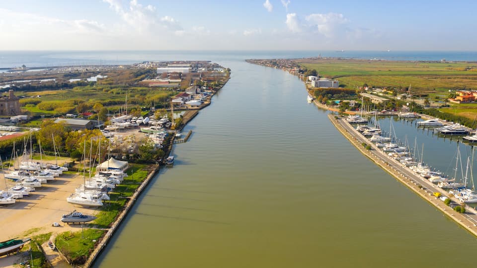 A marina with numerous boats docked along a narrow waterway, surrounded by greenery and industrial buildings.