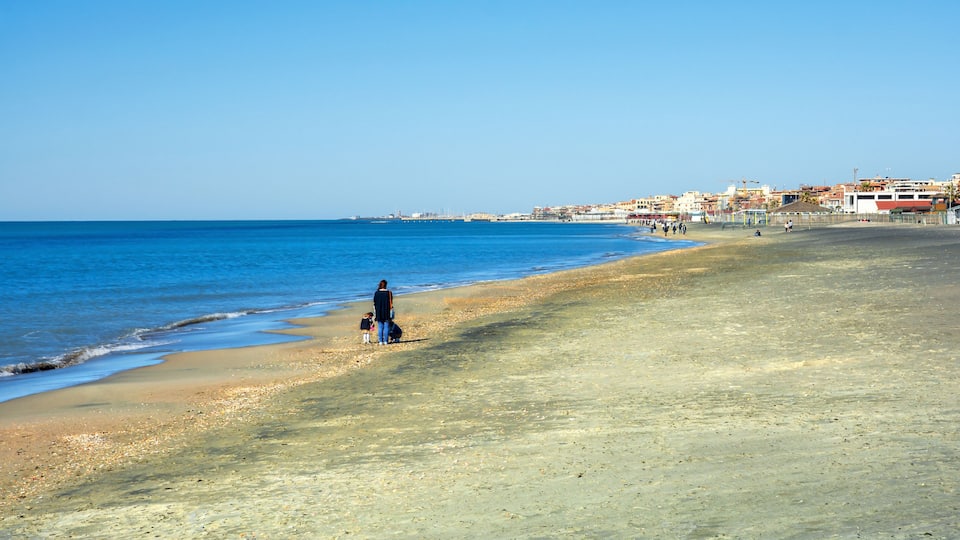 A beach with clear blue water, a sandy shore, and buildings in the background.