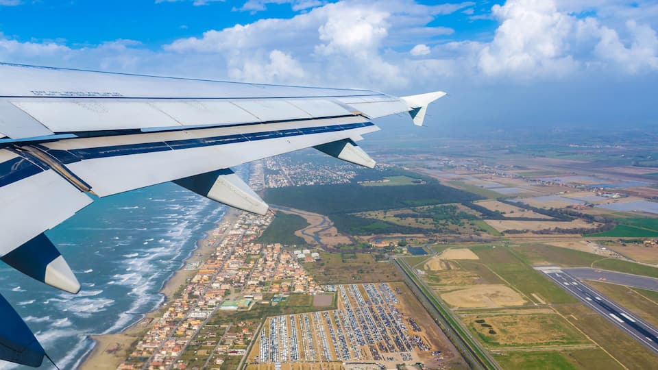 An airplane wing over a coastal city with a large parking lot and a runway.