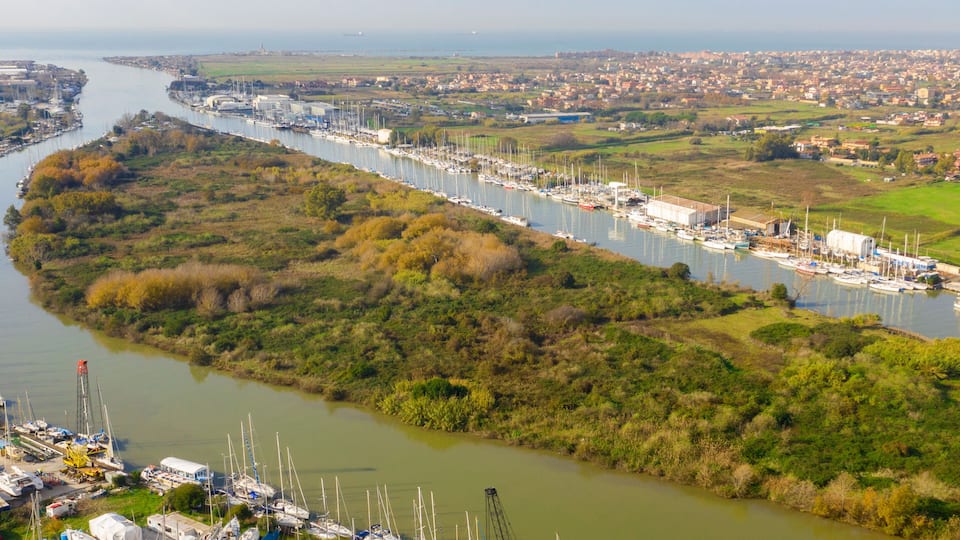 A river with boats docked along its banks, surrounded by greenery and a town in the background.