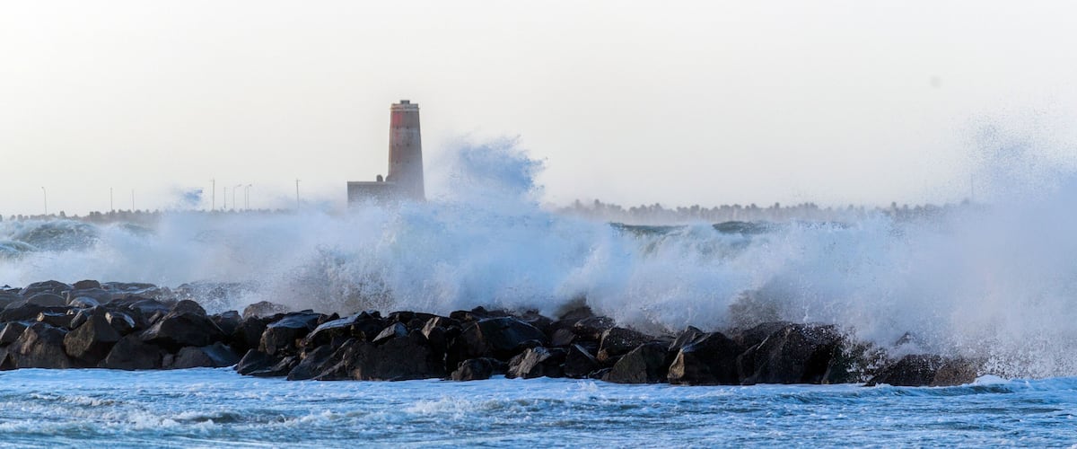 Rough sea waves crashing against a rocky shore with a lighthouse in the background.