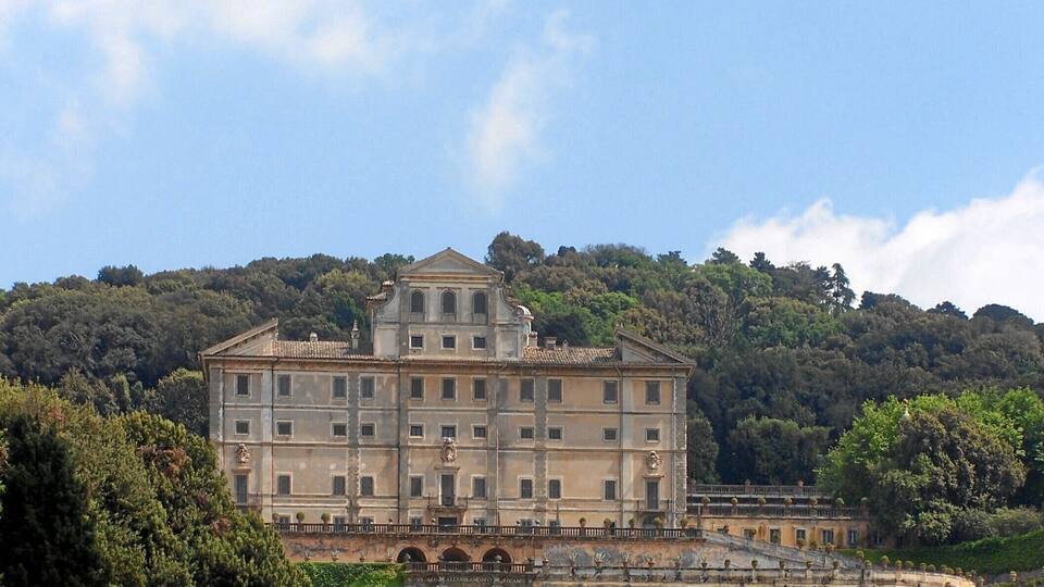 A large, historic building with a symmetrical facade, surrounded by lush greenery and a hilly backdrop.