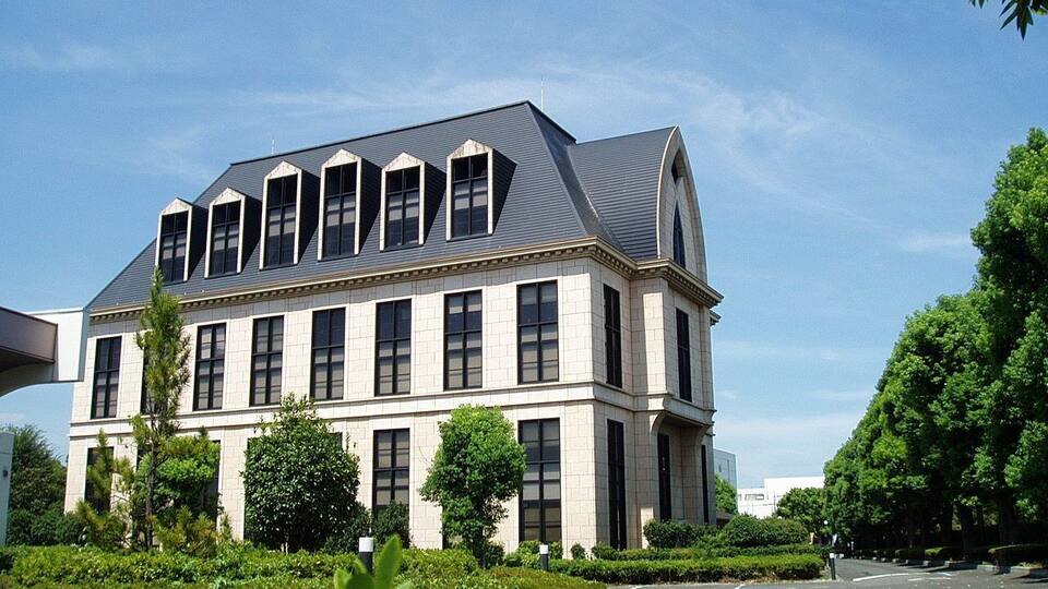 A large, multi-story building with a grey roof and beige walls, surrounded by well-maintained greenery and a clear sky.