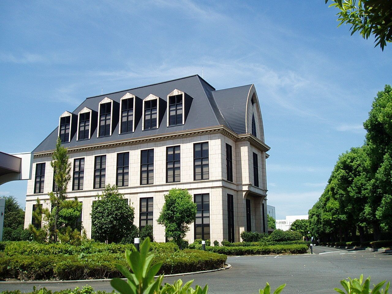 A large, multi-story building with a grey roof and beige walls, surrounded by well-maintained greenery and a clear sky.