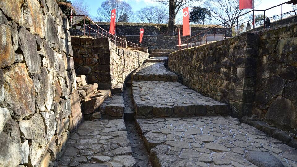 A stone pathway flanked by stone walls, leading up a hill with red flags.