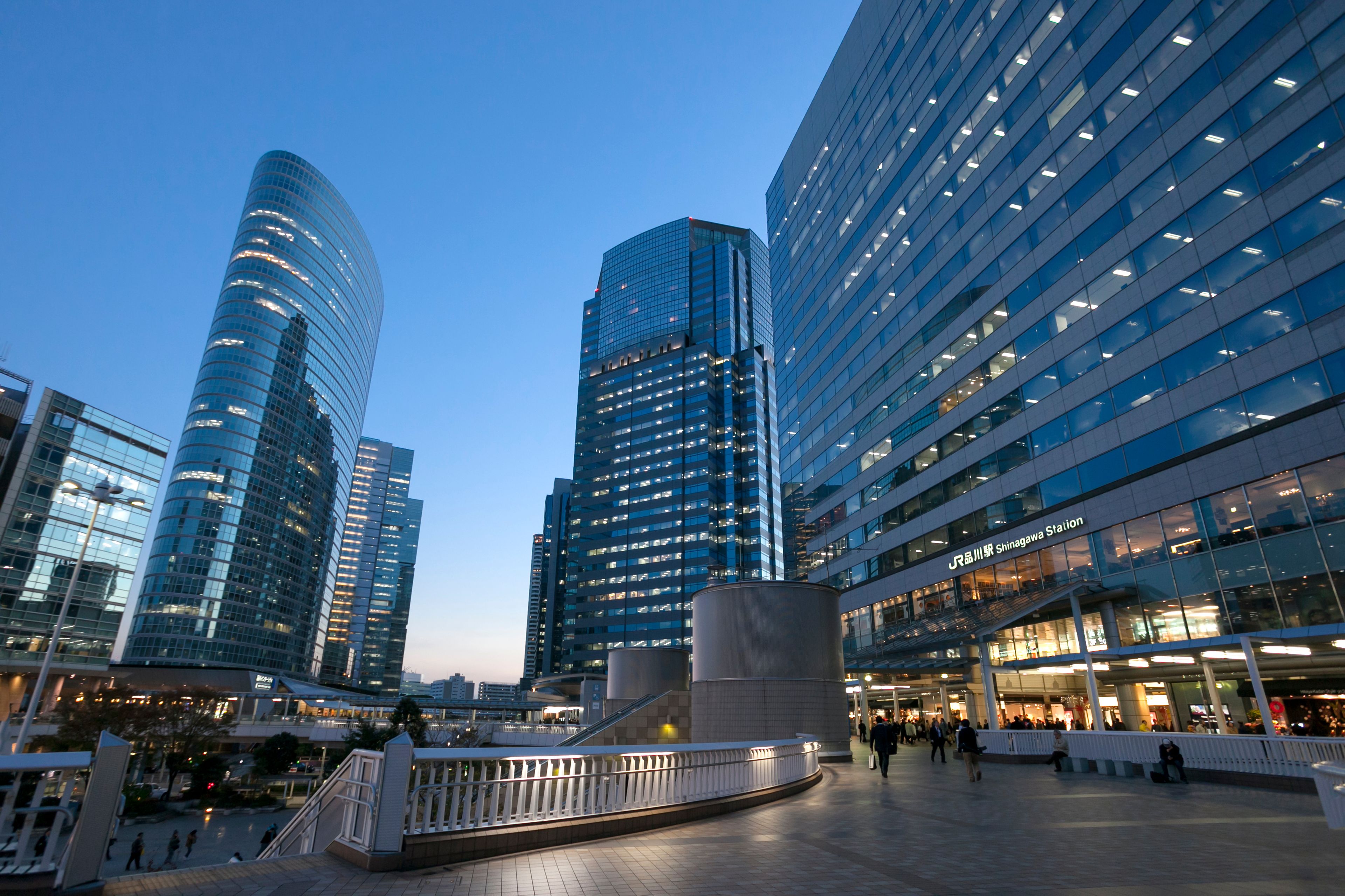A modern urban plaza with high-rise buildings, a curved glass structure, and a pedestrian walkway.