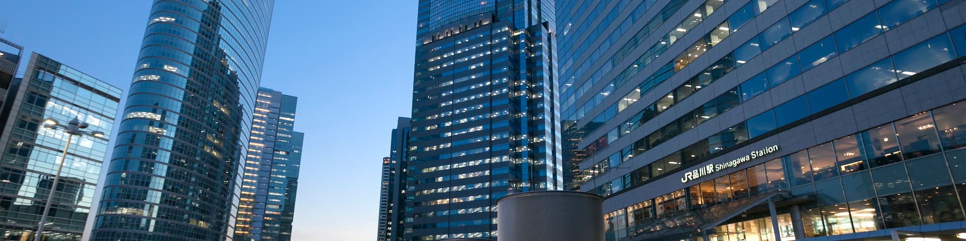 A modern urban plaza with high-rise buildings, a curved glass structure, and a pedestrian walkway.