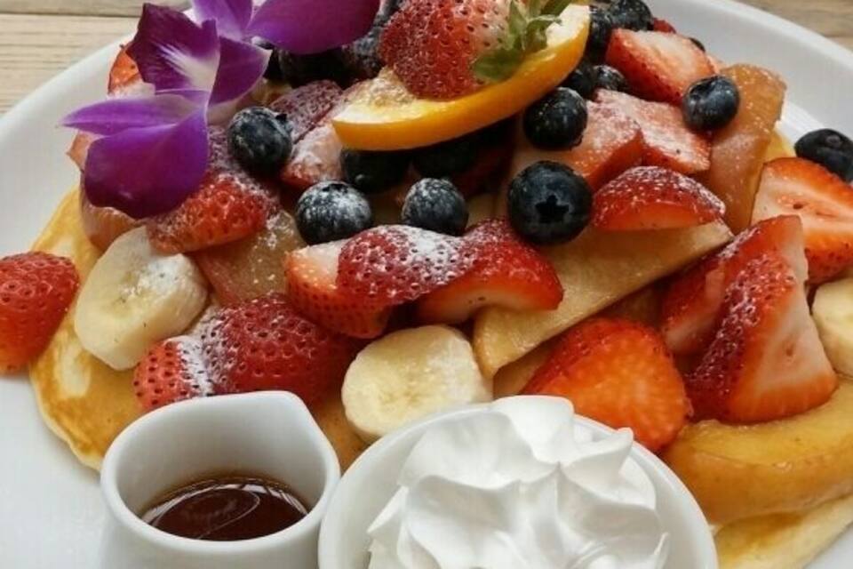 En tallrik med pannkakor, dekorerad med jordgubbar, blåbär, bananskivor och en lila blomma. Det finns två små skålar, en med sirap och en med vispad grädde.