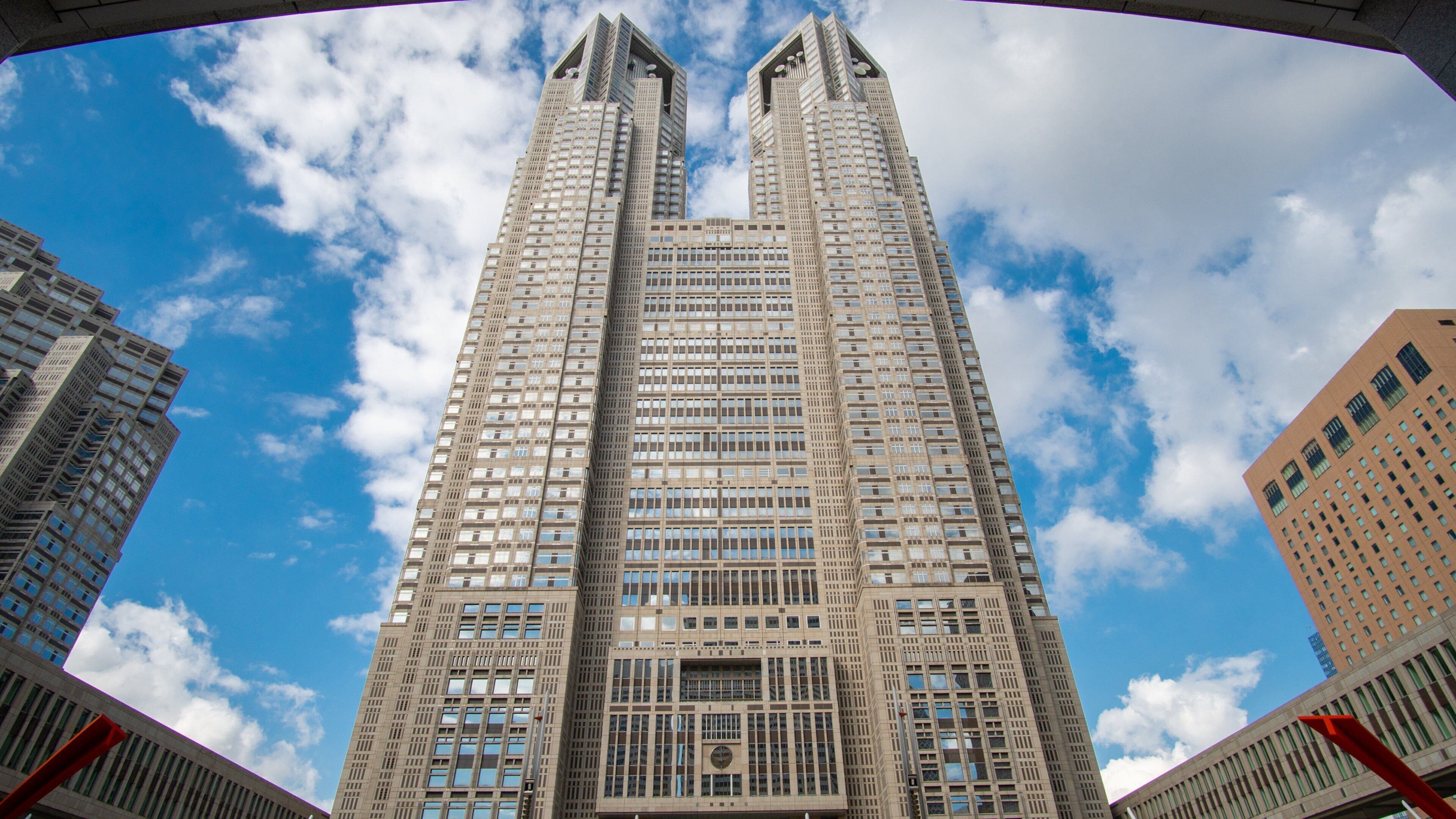 A tall, modern skyscraper with a unique architectural design, surrounded by other buildings and a clear blue sky with clouds.