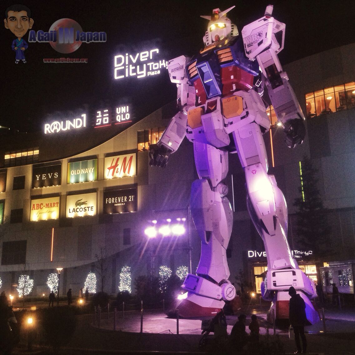 Uma grande estátua de robô em frente a um edifício com letreiros luminosos para lojas como YEVS, Old Navy, H&M e Lacoste.
