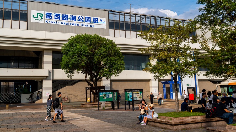 A modern building with signage in Japanese, a tree-lined plaza, and people walking and sitting.