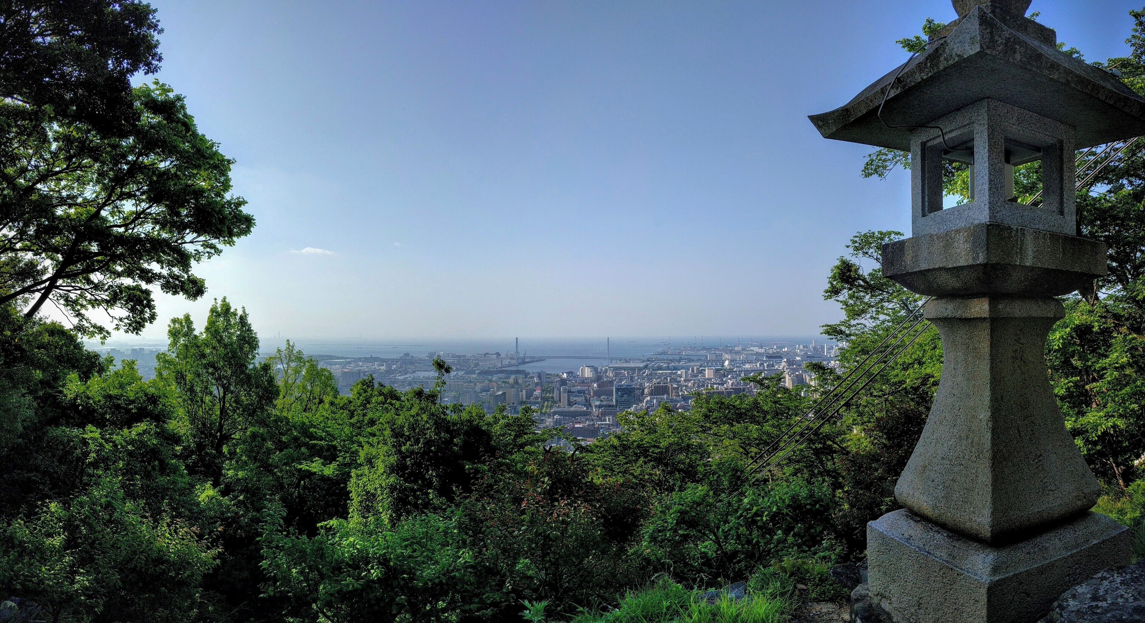A stone lantern amidst lush greenery, overlooking a cityscape with a bridge in the distance.