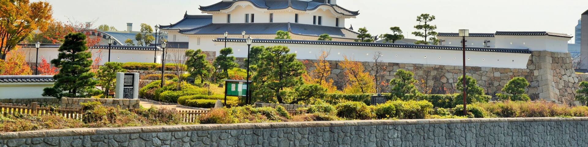 A traditional Japanese castle with multiple tiers, surrounded by a stone wall and lush greenery.