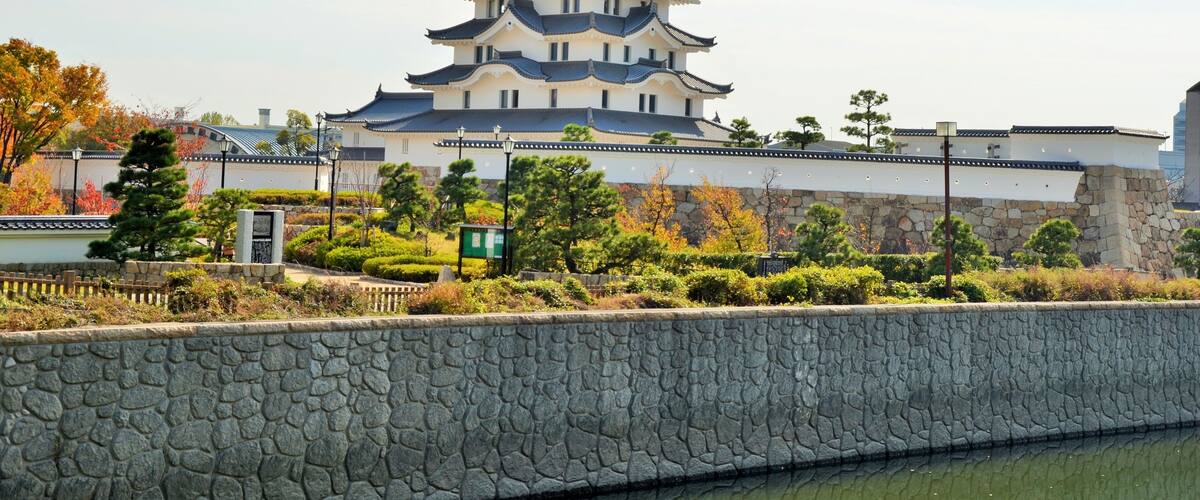 A traditional Japanese castle with multiple tiers, surrounded by a stone wall and lush greenery.