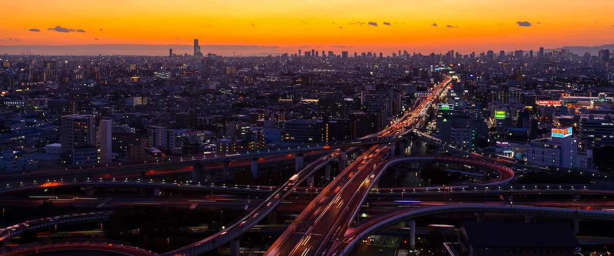 A cityscape at sunset with a prominent highway and dense urban buildings.