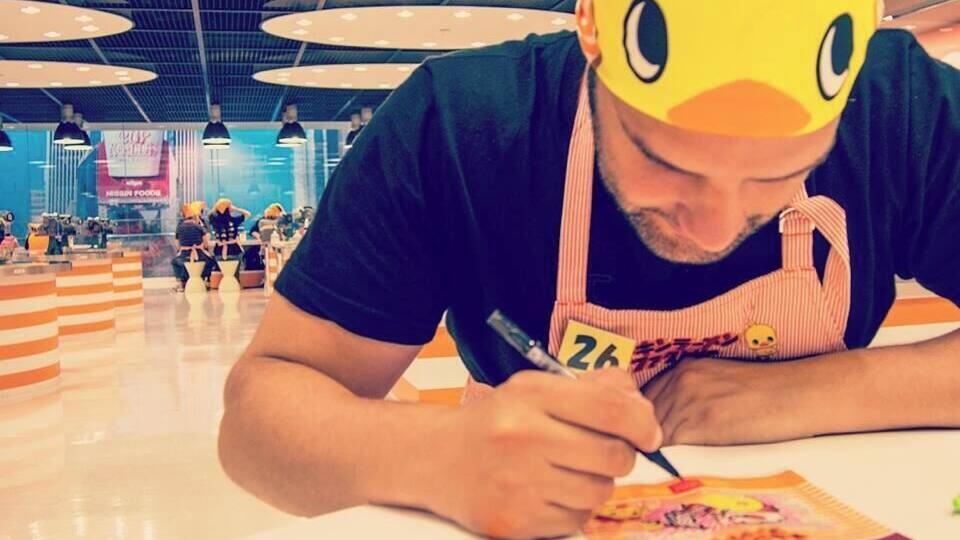A person wearing a yellow duck costume, signing autographs at a table in a brightly lit indoor space with a high ceiling and circular light fixtures.