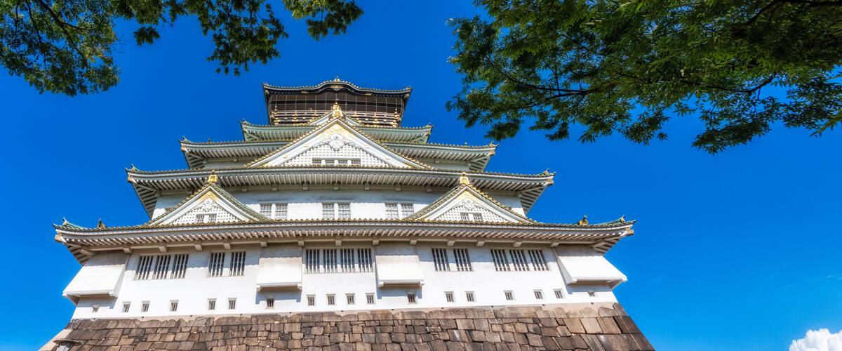 A traditional Japanese castle with multiple tiers, white walls, and golden accents, surrounded by clear blue sky and green foliage.