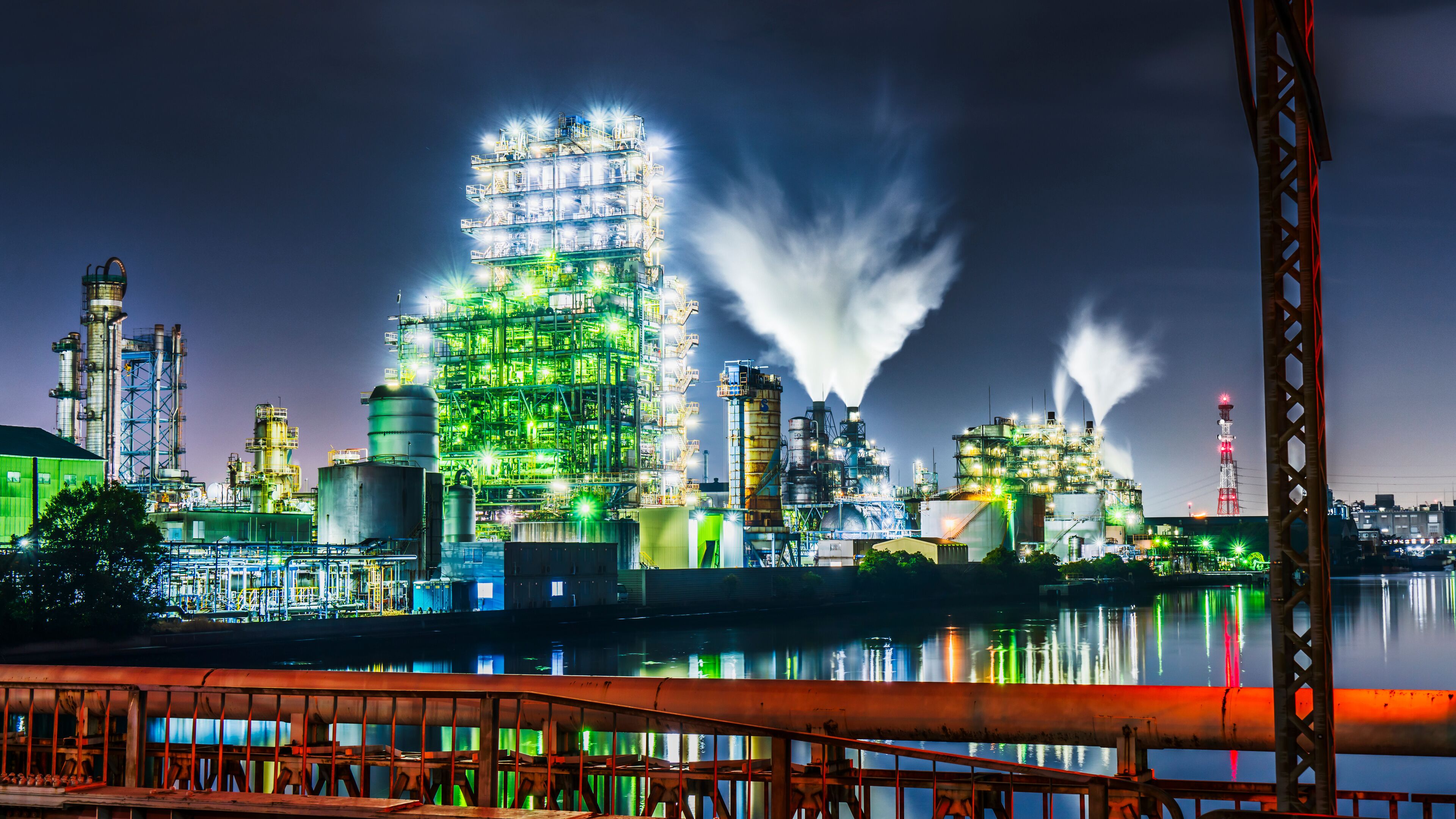 A brightly lit industrial complex with multiple tall structures and smoke plumes against a night sky.