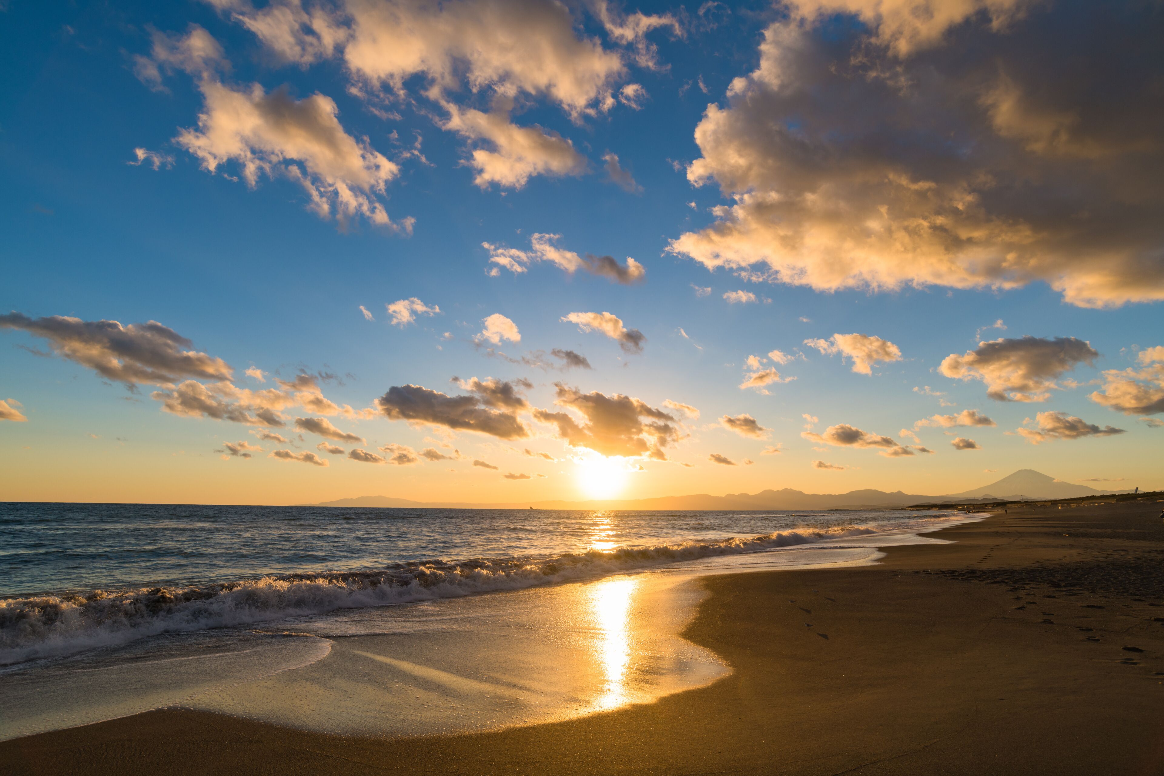 A beach with waves, a sunset, and a mountain in the distance.