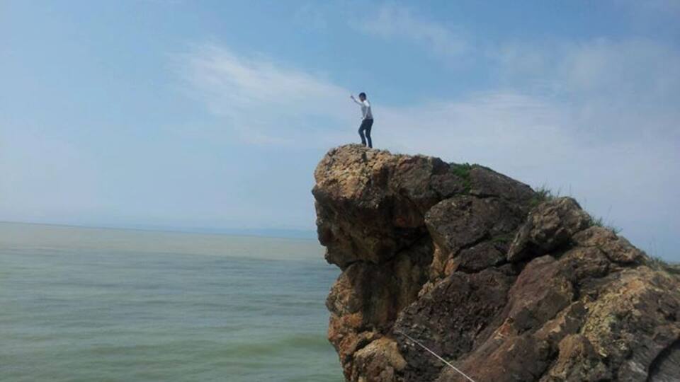 A person standing on a rocky cliff edge with a rope, overlooking a body of water.