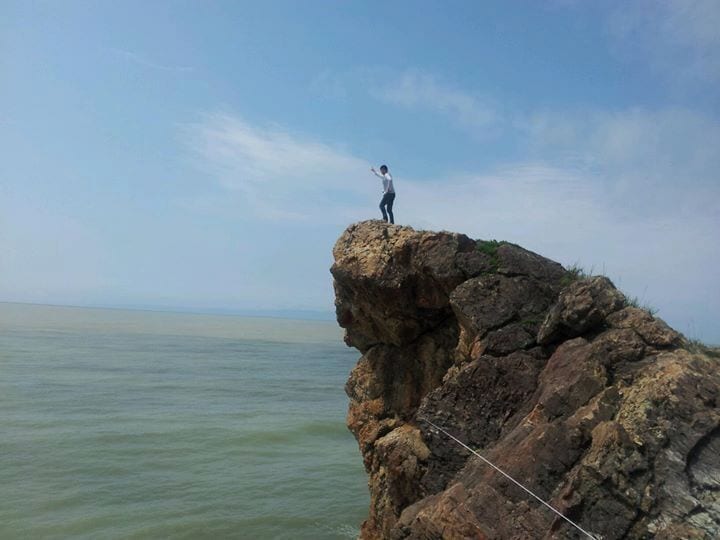 A person standing on a rocky cliff edge with a rope, overlooking a body of water.