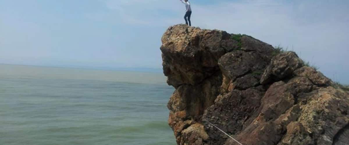 A person standing on a rocky cliff edge with a rope, overlooking a body of water.