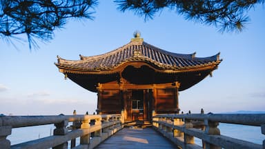 A traditional Chinese pavilion with a tiled roof, situated on a stone bridge over water, with mountains in the background.