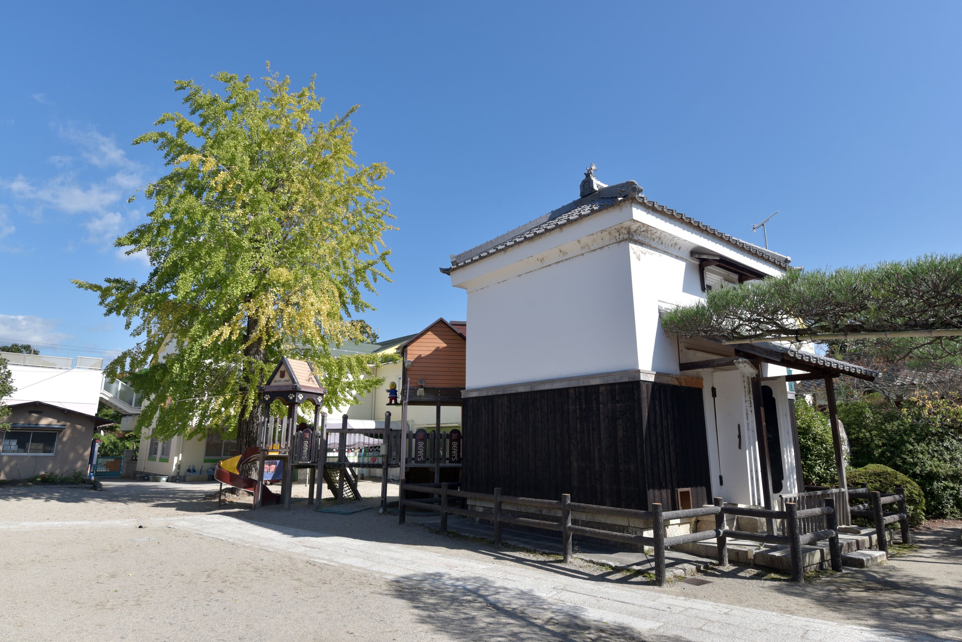 Edificio japonés tradicional con fachada blanca y detalles de madera oscura, rodeado por una cerca de madera y un árbol grande.