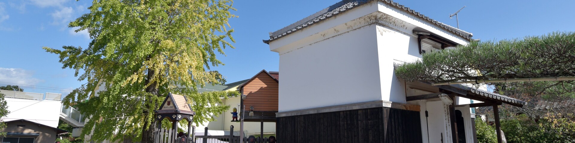 Edificio japonés tradicional con fachada blanca y detalles de madera oscura, rodeado por una cerca de madera y un árbol grande.