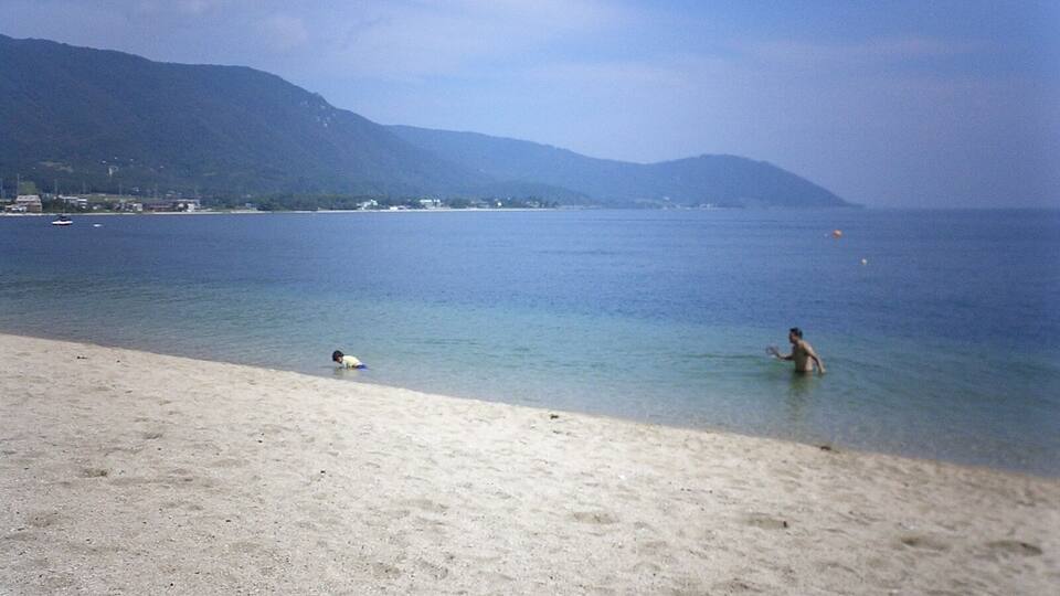 A beach with clear water, a person in the water, and mountains in the background.