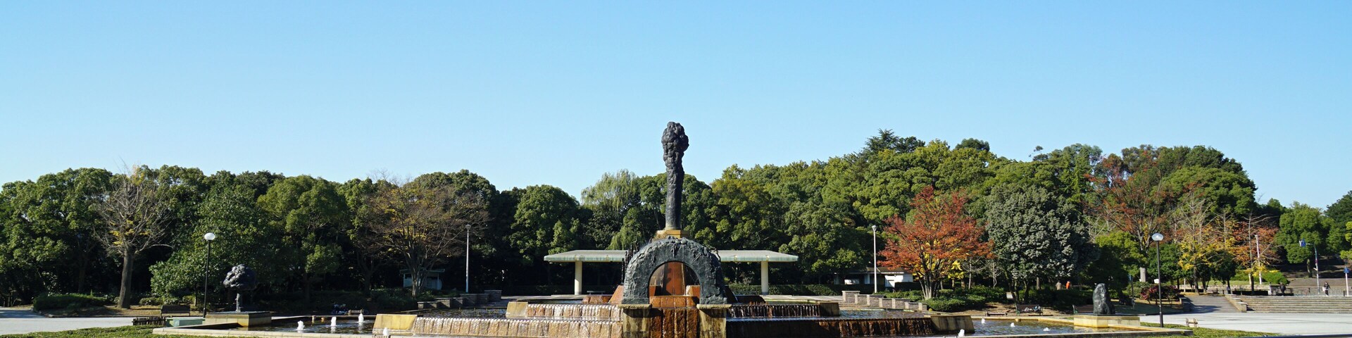 A park with a central fountain, stone pathways, and a backdrop of trees and a clear sky.