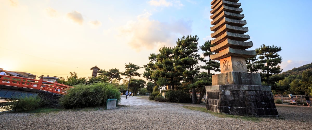 A pagoda-style tower with a stepped design, surrounded by gravel, trees, and a red bridge.
