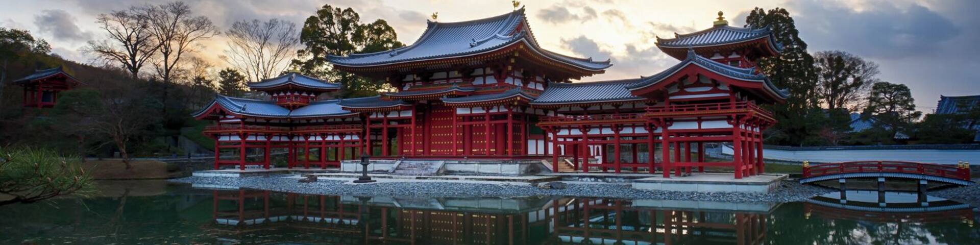 A traditional Japanese temple with red and white buildings reflected in a calm pond, surrounded by trees and under a cloudy sky.