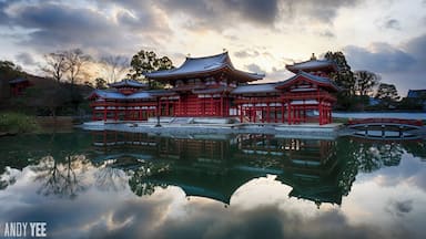 Um templo japonês tradicional com edifícios vermelhos e brancos refletidos num lago calmo, rodeado por árvores sob um céu nublado.