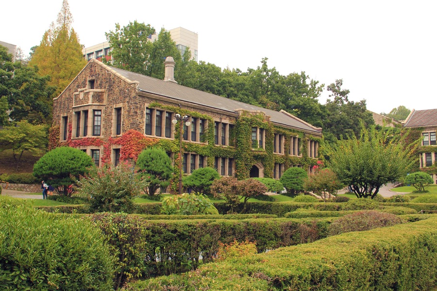 A historic stone building with ivy, surrounded by well-manicured gardens and trees.