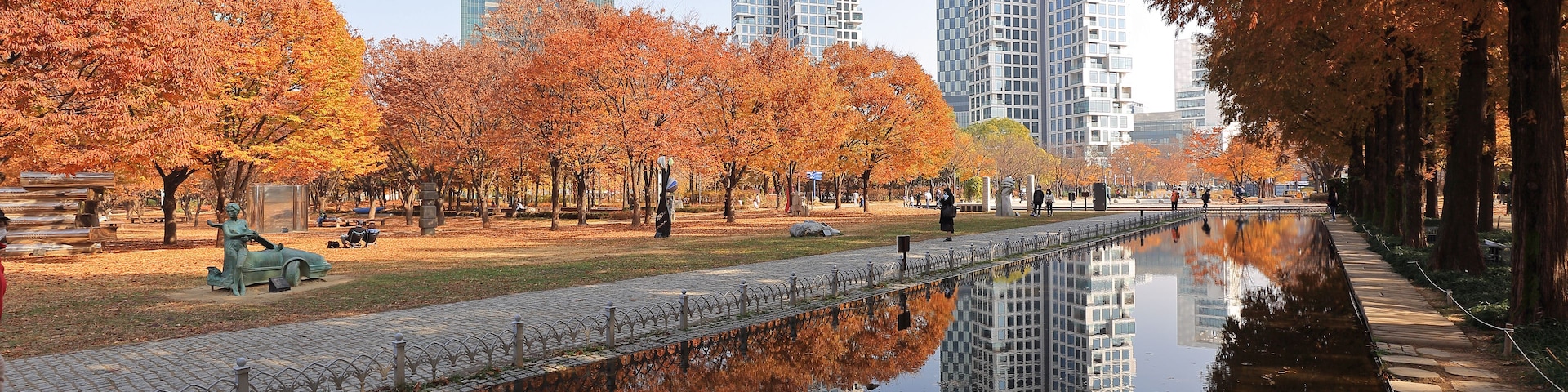 Um parque com árvores de outono, um canal de água reflexiva e edifícios altos e modernos ao fundo.